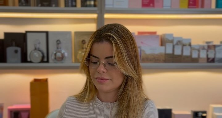 Carol atendendo na loja da Dom Imports, com as prateleiras de perfumes ao fundo